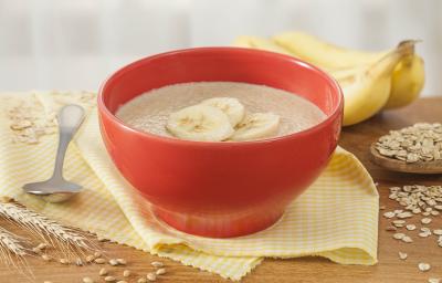 Fotografia em tons de amarelo e vermelho de uma bancada de madeira com um paninho amarelo e um recipiente redondo vermelho, sobre ele o mingau de farinha láctea com fatias de banana. Ao fundo duas bananas e aveias espalhadas pela bancada.