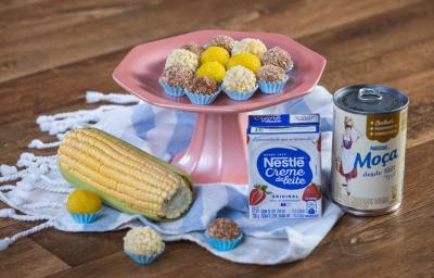 Foto em tons bem coloridos da receita de brigadeiro de milho em várias porções servidas sobre uma base alta cor de rosa com um paninho azul embaixo. Ao lado, há uma espiga de milho, uma caixinha de creme de leite e uma lata de leite moça Foto em tons bem coloridos da receita de brigadeiro de milho em várias porções servidas sobre uma base alta cor de rosa com um paninho azul embaixo. Ao lado, há uma espiga de milho, uma caixinha de creme de leite e uma lata de leite moça