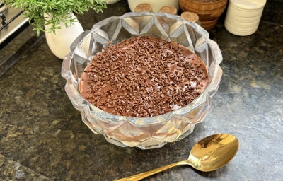 Fotografia mostra um recipiente de vidro redondo com uma cobertura de chocolate e granulado por cima sobre uma bancada preta.