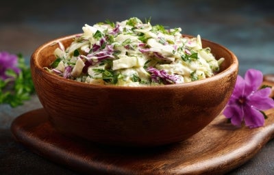 Fotografia de uma salada colorida em uma tigela de madeira, com repolho verde e roxo, ervas frescas e um molho cremoso.