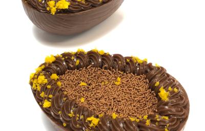 Foto em tons de branco, marrom claro, escuro e laranja. Na foto é possível ver uma bancada branca com duas cascas de ovos recheadas com brigadeiro, chocolate granulado e migalhas de bolo de cenoura Foto em tons de branco, marrom claro, escuro e laranja. Na foto é possível ver uma bancada branca com duas cascas de ovos recheadas com brigadeiro, chocolate granulado e migalhas de bolo de cenoura