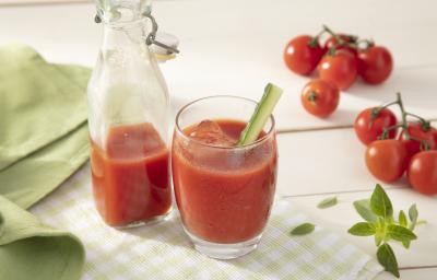 Fotografia em tons de vermelho em uma bancada de madeira clara, um pano verde liso e outro xadrez, um copo de vidro com o suco de tomate e uma jarra com mais suco ao centro da foto. Ao lado, tomates pequenos para decorar a mesa. Fotografia em tons de vermelho em uma bancada de madeira clara, um pano verde liso e outro xadrez, um copo de vidro com o suco de tomate e uma jarra com mais suco ao centro da foto. Ao lado, tomates pequenos para decorar a mesa.