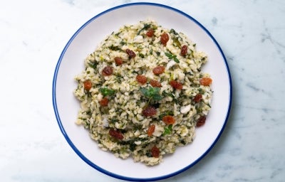 Visto de cima, prato redondo branco com porção de arroz cremoso verde e uvas-passas. Decorado com uma folha de manjericão.