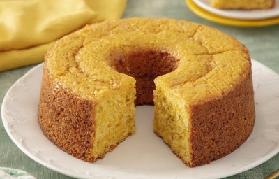 Bolo de milho dourado e fofinho, inteiro com fatia cortada, servido em prato branco sobre mesa rústica.