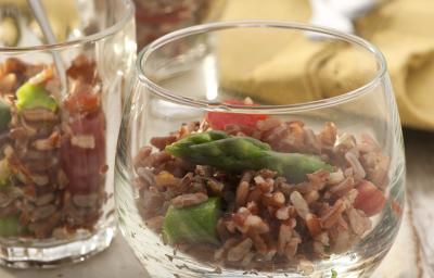 Fotografia em tons de marrom de uma bancada de madeira com uma tábua quadrada marrom, sobre ela um recipiente de vidro com salada de arroz vermelho. Ao fundo um paninho amarelo.