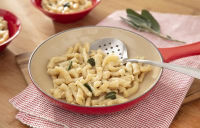 Foto da receita de Spatzle. Observa-se uma frigideira grande sobre um pano de prato quadriculado vermelho. A massa está dentro da frigideira com folhas de manjericão. Foto da receita de Spatzle. Observa-se uma frigideira grande sobre um pano de prato quadriculado vermelho. A massa está dentro da frigideira com folhas de manjericão.