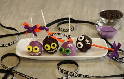 Imagem de um prato branco com 4 bolinhas decoradas com palitos brancos. A cena está decorada com uma fita preta com caveiras brancas e dois potes com chocolate roxo e granulados pretos. Imagem de um prato branco com 4 bolinhas decoradas com palitos brancos. A cena está decorada com uma fita preta com caveiras brancas e dois potes com chocolate roxo e granulados pretos.