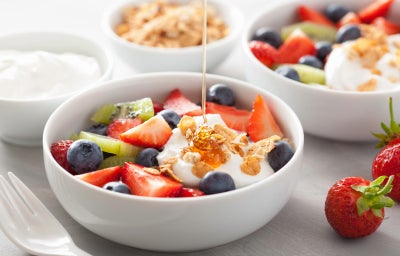 Fotografia em tons de branco, com três refratários brancos fundos no canto e no centro outro refratário com diversas frutas, iogurte e uma calda de mel caindo. No canto inferior direito, dois morangos inteiros.