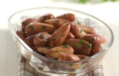 Fotografia em tons de marrom, com pote de vidro ao centro com pinhão sauteado salpicado de salsa, sobre guardanapo quadriculado nas cores branca e marrom, ao fundo ramo de salsa, tudo sobre bancada bege. Fotografia em tons de marrom, com pote de vidro ao centro com pinhão sauteado salpicado de salsa, sobre guardanapo quadriculado nas cores branca e marrom, ao fundo ramo de salsa, tudo sobre bancada bege.