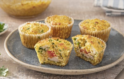 Fotografia em tons claros com pequenas tortinhas de frango ao centro. As tortinhas de frango são feitas em formas de cupcake com frango desfiado, tomate e ervas frescas
