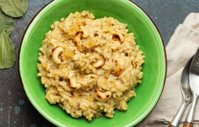 Fotografia vista de cima de um arroz crocante com castanha de caju dentro de um recipiente fundo verde, o qual está sobre uma mesa escura preta. Ao lado do recipiente tem um paninho e talheres por cima.
