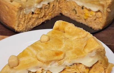 Fotografia de uma fatia de empadão de frango sobre um prato raso branco de vidro. No fundo tem o resto da torta quase inteira sobre o fundo da forma removível.