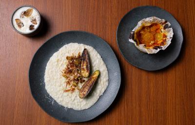 Fotografia vista de cima de três pratos pratos pretos com receitas diferentes, sendo elas: Fonduta de Gruyère com Cogumelos, Risotto de Brie com Abobrinha e Torta de Queijo com Compota de Frutas Amarelas. Os prato estão sobre uma bancada de madeira.