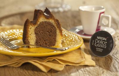 Fotografia em tons de marrom e amarelo de uma bancada de madeira com um paninho amarelo, sobre ele um prato redondo amarelo com uma fatia do bolo e um garfo. Ao fundo uma xícara branca com detalhes vermelhos e uma capsula de Dolce Gusto. Fotografia em tons de marrom e amarelo de uma bancada de madeira com um paninho amarelo, sobre ele um prato redondo amarelo com uma fatia do bolo e um garfo. Ao fundo uma xícara branca com detalhes vermelhos e uma capsula de Dolce Gusto.