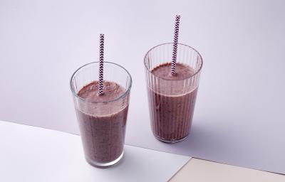 Fotografia em tons de roxo, em uma bancada de madeira, uma toalha lilás, um pano branco, dois copos de vidro altos com a vitamina de frutas vermelhas dentro deles, e um canudinho roxo dentro de cada copo.