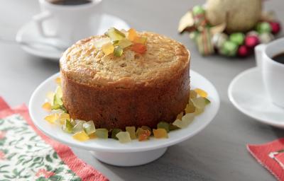 Fotografia em tons de amarelo em uma bancada de madeira de cor cinza. Ao centro, uma boleira contendo o panettone com frutas cristalizadas e uma toalha florida ao lado. Ao fundo, um pires com uma xícara branca de café e alguns enfeites natalinos.