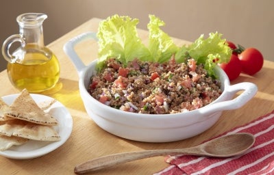 Fotografia em tons claros de um recipiente branco e fundo de vidro com uma tabule com folhas de alface. Ao redor do recipiente tem azeite, tomates pequenos, colher de madeira e pães sírios em um mini pratinho branco sobre uma mesa de madeira. Fotografia em tons claros de um recipiente branco e fundo de vidro com uma tabule com folhas de alface. Ao redor do recipiente tem azeite, tomates pequenos, colher de madeira e pães sírios em um mini pratinho branco sobre uma mesa de madeira.
