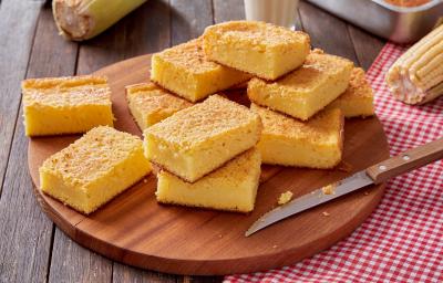 Foto da receita de bolo de milho cremoso, fatiado em pedaços sobre uma tábua de madeira sobre um pano quadriculado vermelho e branco. Na mesa há uma faca e espigas de milho Foto da receita de bolo de milho cremoso, fatiado em pedaços sobre uma tábua de madeira sobre um pano quadriculado vermelho e branco. Na mesa há uma faca e espigas de milho