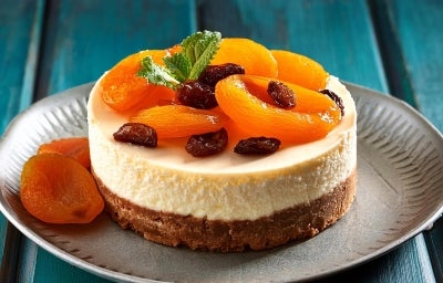 Fotografia de uma mini torta inteira sobre um prato cinza pequeno sore uma mesa de madeira azul. Sobre a torta tem damascos e frutas secas. Fotografia de uma mini torta inteira sobre um prato cinza pequeno sore uma mesa de madeira azul. Sobre a torta tem damascos e frutas secas.