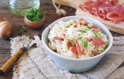 Fotografia em tons claros de um refratário branco pequeno e fundo sobre uma toalha de mesa, com arroz cremoso, peito de peru, lascas de presunto de Parma e cebolinha por cima. Ao fundo, fatias do presunto sobre uma tábua de madeira e algumas decorações. Fotografia em tons claros de um refratário branco pequeno e fundo sobre uma toalha de mesa, com arroz cremoso, peito de peru, lascas de presunto de Parma e cebolinha por cima. Ao fundo, fatias do presunto sobre uma tábua de madeira e algumas decorações.