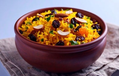 Tigela de porcelana redonda com porção de arroz amarelo e soltinho com amêndoas e uvas-passas pretas, decorado com folhas de cheiro-verde.