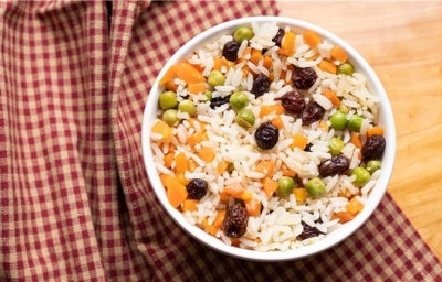 Em cima de uma mesa de madeira, há um pote branco  com uma porção colorida de arroz branco com uvas-passas, cenouras e ervilhas.