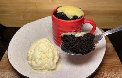 Fotografia em tons claros de um prato branco de vidro sobre uma tábua de madeira. Por cima do prato tem uma caneca vermelha com bolo de micro-ondas, cobertura de limão e, ao lado, uma bola de sorvete de creme e uma colher segurando o bolo. Fotografia em tons claros de um prato branco de vidro sobre uma tábua de madeira. Por cima do prato tem uma caneca vermelha com bolo de micro-ondas, cobertura de limão e, ao lado, uma bola de sorvete de creme e uma colher segurando o bolo.