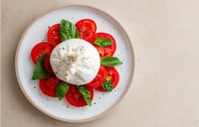 Fotografia vista de cima em tons claros de uma mesa de tom bege. Sobre a mesa tem um prato de cerâmica branco com uma burrata ao centro e rodelas de tomate em volta. Entre os tomates tem folhas de manjericão, sobre a burrata tem molho de manjericão.