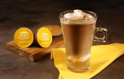 Fotografia em tons de amarelo em uma bancada de madeira escura, um paninho amarelo, uma xícara de vidro alta com o Latte Macchiato Dolce Gusto dentro, com doce de leite e decorado com chantilly. Ao fundo, cápsulas do café e um potinho com doce de leite. Fotografia em tons de amarelo em uma bancada de madeira escura, um paninho amarelo, uma xícara de vidro alta com o Latte Macchiato Dolce Gusto dentro, com doce de leite e decorado com chantilly. Ao fundo, cápsulas do café e um potinho com doce de leite.