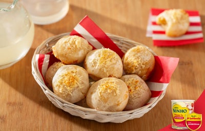 Pãezinhos de queijo dourados dispostos em cesta com papel vermelho e branco, sobre mesa de madeira, com bebida ao fundo.