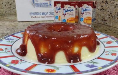 Imagem de um Pudim Romeu e Julieta sobre um prato colorido, numa bancada de cozinha com algumas embalagens do produto Moça Pra Toda a Família ao fundo
