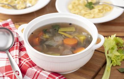 Fotografia em tons de vermelho em uma bancada de madeira, um pano xadrez vermelho, uma panela branca funda com alças laterais com o caldo de legumes dentro dela. Ao lado, uma concha e dois pratos brancos fundo com sopa de capelleti com o caldo de legumes. Fotografia em tons de vermelho em uma bancada de madeira, um pano xadrez vermelho, uma panela branca funda com alças laterais com o caldo de legumes dentro dela. Ao lado, uma concha e dois pratos brancos fundo com sopa de capelleti com o caldo de legumes.