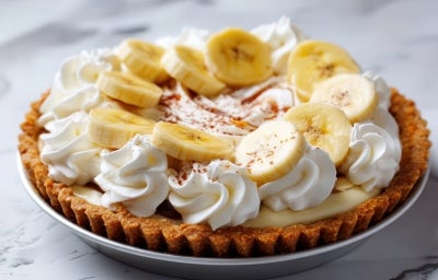 Fotografia em tons claros de uma banoffee inteira e completa dentro de um recipiente meio fundo sobre uma mesa de mármore.