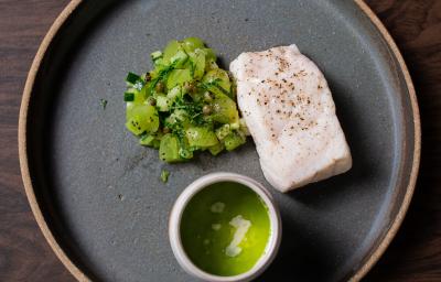 Fotografia vista de cima de um prato raso grande preto com uma posta de robalo no vapor com molho de leite de coco, ervas e salada de frutas verdes. O prato está sobre uma bancada de madeira.