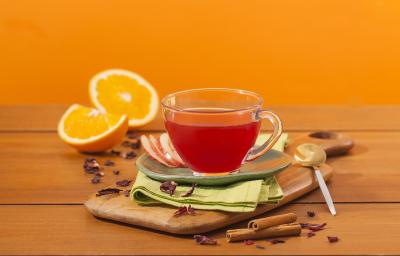 Fotografia em tons de laranja em uma bancada de madeira e um fundo laranja. Ao centro, uma tábua de madeira, um paninho verde e uma xícara de vidro com o chá de hibisco dentro dele. Ao fundo, laranja e canela em pau espalhados pela bancada. Fotografia em tons de laranja em uma bancada de madeira e um fundo laranja. Ao centro, uma tábua de madeira, um paninho verde e uma xícara de vidro com o chá de hibisco dentro dele. Ao fundo, laranja e canela em pau espalhados pela bancada.