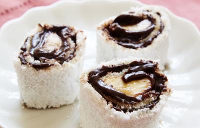 Fotografia em tons de vermelho em uma bancada de madeira clara, um pano vermelho, um prato branco redondo com três rolinhos de tapiocas com banana e chocolate.