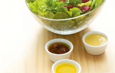 Fotografia em tons de verde em uma mesa de madeira clara com um bowl de vidro com salada de alface e rúcula e ao lado, quatro potinhos brancos com os molhos de morango, mostarda, hortelã e gengibre.