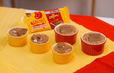 Cocadas cremosas em ramequins coloridos, cobertas com chocolate recheado, com embalagem Garoto ao fundo.