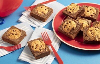 Fotografia em tons de azul e vermelho de uma bancada azul com paninhos azul claro com listras brancas e sobre eles pedaços de brownie com cookies e garfos vermelhos. Ao lado um prato vermelho e sobre eles pedaços de brownie com cookies.