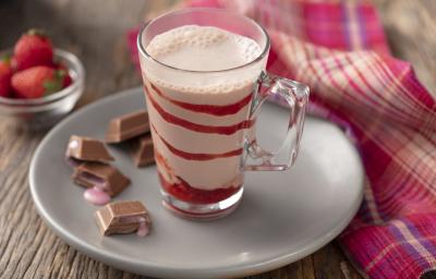 fotografia em tons de marrom, branco, vermelho e rosa de uma bancada marrom vista de frente. Contém um prato redondo branco, com um copo transparente que contém a bebida de morango, e ao lado cubos de chocolate com recheio de morango. fotografia em tons de marrom, branco, vermelho e rosa de uma bancada marrom vista de frente. Contém um prato redondo branco, com um copo transparente que contém a bebida de morango, e ao lado cubos de chocolate com recheio de morango.