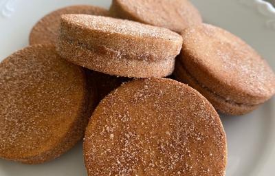 Fotografia em tons de marrom com um prato branco ao centro. Dentro do prato existe algumas unidades de biscoitos de cor caramelo recheados com doce de leite e cobertos com açúcar. Fotografia em tons de marrom com um prato branco ao centro. Dentro do prato existe algumas unidades de biscoitos de cor caramelo recheados com doce de leite e cobertos com açúcar.