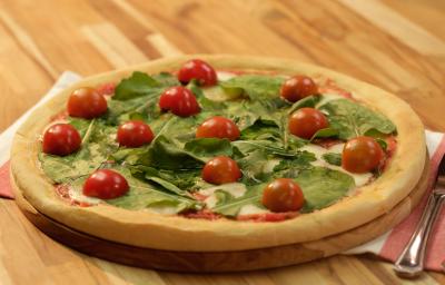 Fotografia em tons de dourado, verde e vermelho, ao centro pizza com folhas verdes e tomate-cereja sobre tábua redonda e guardanapo branco e vermelho, com talher ao lado, tudo sobre bancada de madeira em tons marrom.