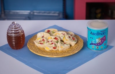 Fotografia em tons claros de um pano azul e quadrado sobre uma mesa de tom claro. Sobre esse paninho tem um prato desenhado amarelo, de vidro, com os sushis de frutas. Ao lado do prato tem a lata de ascenda e um vidro com mel.