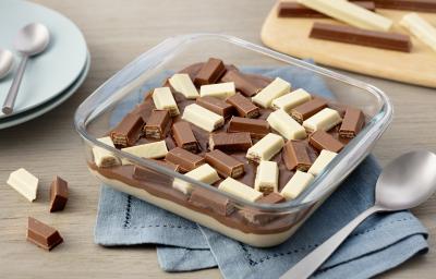 Fotografia em tons de azul em uma bancada de madeira com um pano azul, um refratário de vidro quadrado com o doce de chocolate branco e ao leite dentro, com KitKat ao leite e branco para decorar.