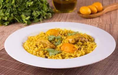 Fotografia em tons de amarelo, em uma bancada de madeira de cor marrom. Ao centro, um prato branco redondo contendo o arroz com frango e pequi. Ao fundo, uma colher de pau contendo alguns pequis e ao lado alguns temperos verdes espalhados. Fotografia em tons de amarelo, em uma bancada de madeira de cor marrom. Ao centro, um prato branco redondo contendo o arroz com frango e pequi. Ao fundo, uma colher de pau contendo alguns pequis e ao lado alguns temperos verdes espalhados.