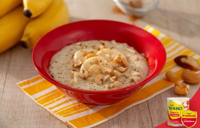 Mingau cremoso servido em tigela vermelha, com banana e castanhas picadas por cima, sobre guardanapo amarelo, em mesa clara.