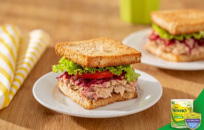 Sanduíche de atum em pão integral, recheado com creme claro, folhas de alface e tomate.