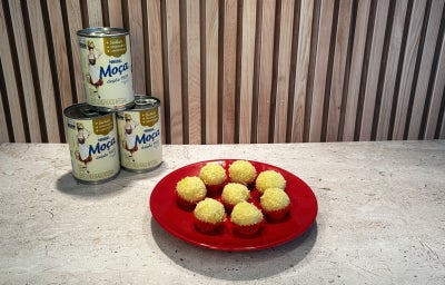 Imagem em tons de bege em um fundo marrom, com Leite Moça ao fundo e ao centro um prato vermelho com os brigadeiros.