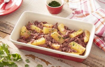 Fotografia em tons de vermelho de uma bancada de madeira, ao centro uma travessa vermelha por fora e branca por dentro com pedaços de mandioca e carne-seca desfiada. Ao redor, folhas de salsa e um paninho quadriculado branco e vermelho. Fotografia em tons de vermelho de uma bancada de madeira, ao centro uma travessa vermelha por fora e branca por dentro com pedaços de mandioca e carne-seca desfiada. Ao redor, folhas de salsa e um paninho quadriculado branco e vermelho.