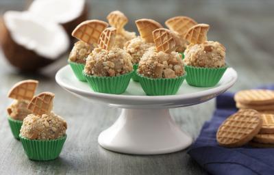 Fotografia em tons de cinza e verde de uma bancada cinza, com um prato branco alto com sete brigadeiros em forminhas verdes com pedaço de biscoito em cima. Ao lado dois brigadeiros, pano azul e bolachas.